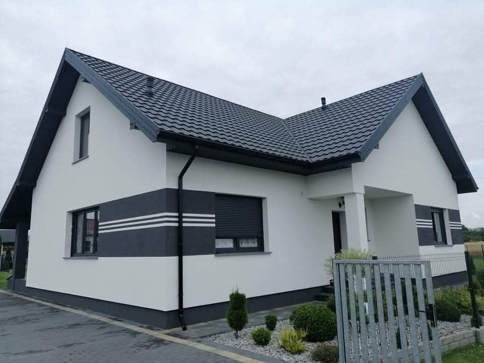 Nowoczesny dom jednorodzinny z białą elewacją, szarym pasem z trzema białymi liniami i ciemną dachówką, widok z zewnątrz na tle pochmurnego nieba.