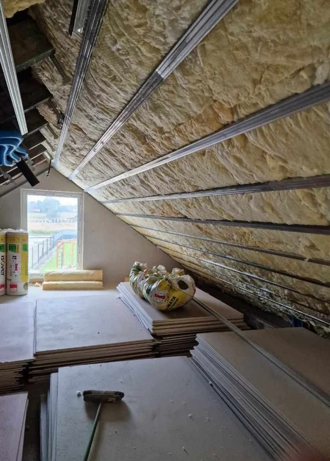 Poddasze w trakcie izolacji: widoczne płyty kartonowo-gipsowe, wełna mineralna między metalowymi profilami, okno dachowe w szczycie.