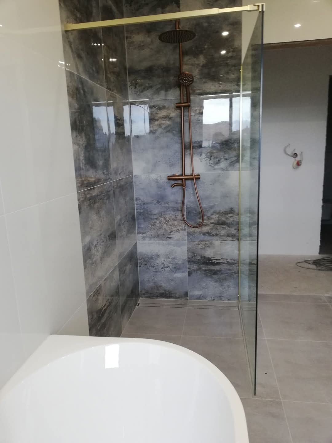 Wykończona łazienka z kabiną prysznicową typu walk-in, deszczownicą w kolorze miedzi i płytkami imitującymi kamień w odcieniach szarości i granatu, widoczna wanna.