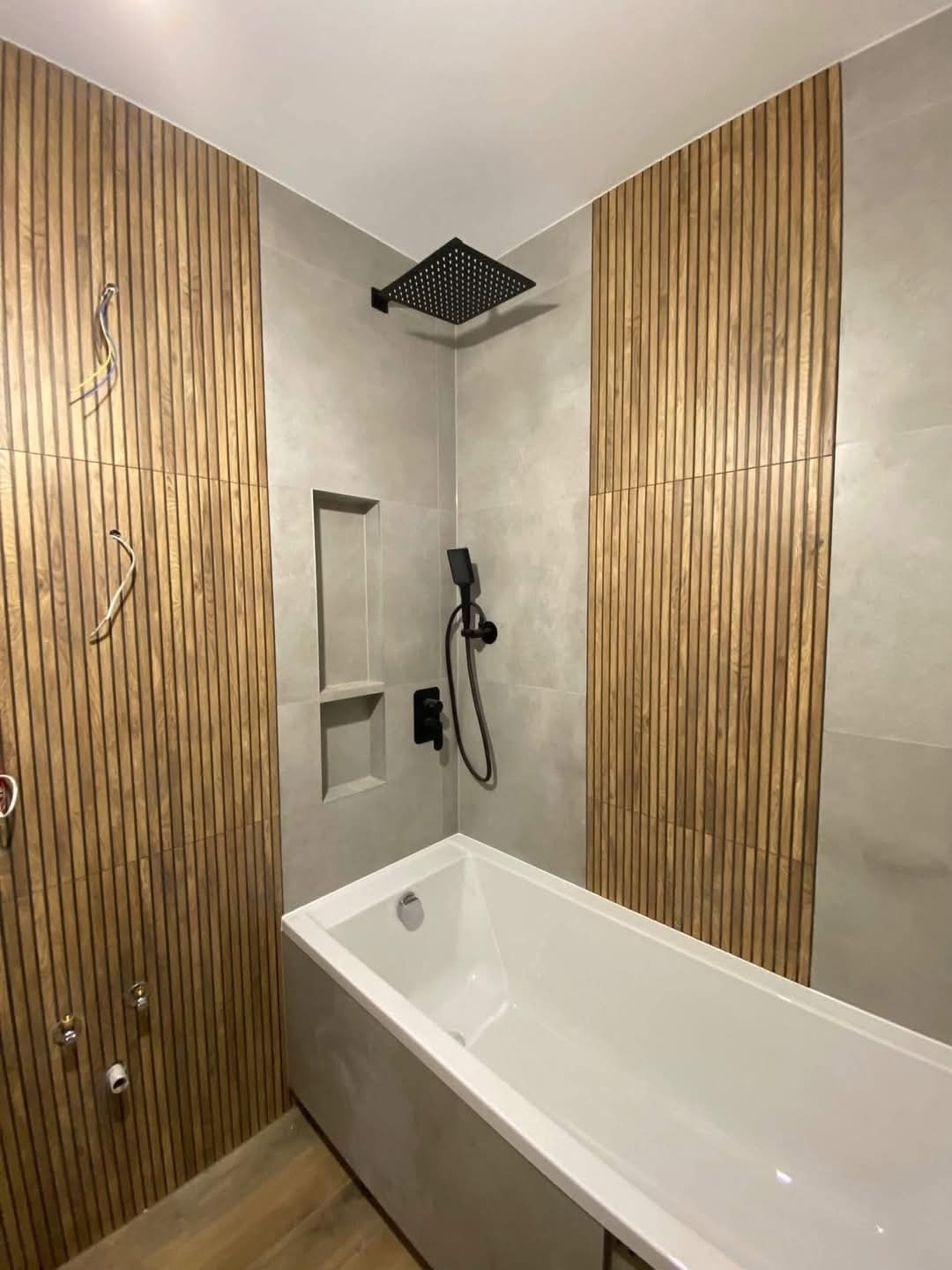 Nowoczesna łazienka z drewnianymi panelami ściennymi, szarą płytką, białą wanną i czarną armaturą, widoczne niedokończone podłączenia instalacji.