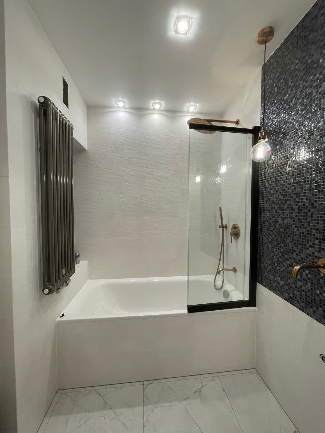 Nowoczesna łazienka z wanną obudowaną płytkami, szklanym parawanem nawannowym z czarną ramą, grzejnikiem drabinkowym w kolorze grafitowym, mozaiką na ścianie i marmurową podłogą.