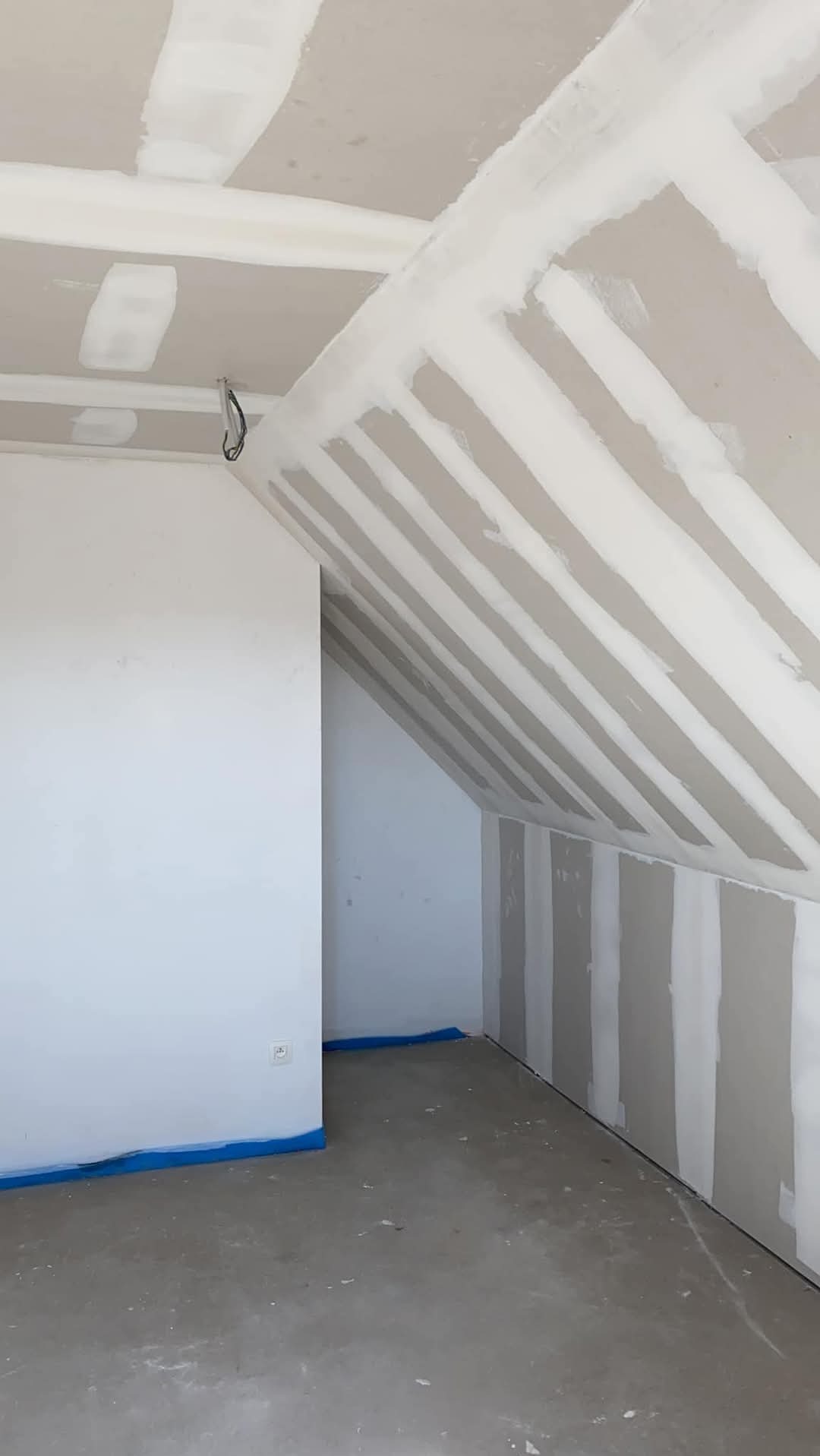 Poddasze w trakcie wykańczania: widoczne płyty kartonowo-gipsowe na skosach dachu i ścianach, łączenia szpachlowane, niebieska folia zabezpieczająca podłogę przy ścianach, surowy beton na podłodze.