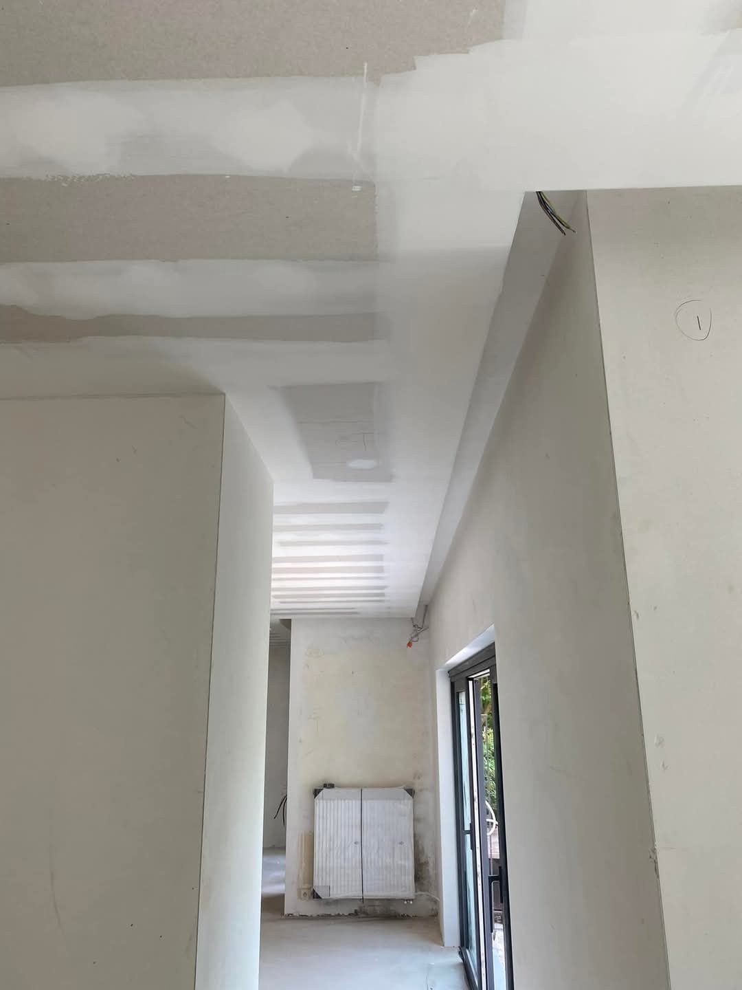 Wnętrze w trakcie prac wykończeniowych: sufit z płyt kartonowo-gipsowych z widocznymi łączeniami, ściany przygotowane pod malowanie, widoczne przewody elektryczne i grzejnik zabezpieczony folią.