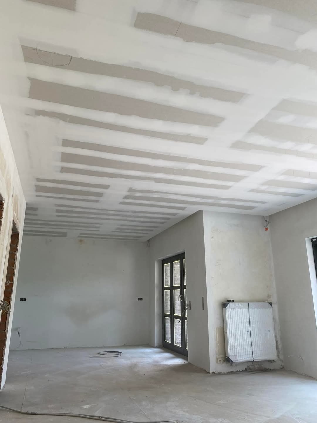 Wnętrze w trakcie prac w miejscowości Włocławek: sufit z płyt kartonowo-gipsowych, częściowo zaszpachlowany, widoczne łączenia płyt. Na ścianach widoczne ślady po instalacjach elektrycznych.