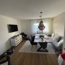 Komfort House Wykończenia Wnętrz Emil Dudziński - Jasne wnętrze jadalni z drewnianym stołem, krzesłami, sofą i komodą. Na podłodze panele. Centralnie umieszczony ozdobny żyrandol. Minimalistyczny wystrój.