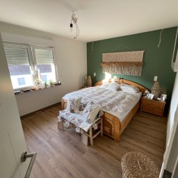 Komfort House Wykończenia Wnętrz Emil Dudziński - Jasna sypialnia z drewnianym łóżkiem, zieloną ścianą i dekoracyjnym makramem. Widoczna podłoga z paneli i częściowo otwarte drzwi.