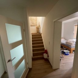 Komfort House Wykończenia Wnętrz Emil Dudziński - Wnętrze domu po remoncie: widok na schody z dekoracją, drzwi z mlecznym szkłem i fragment sypialni z łóżkiem i wiadrem z farbą. Jasne ściany, drewniane schody i podłoga.