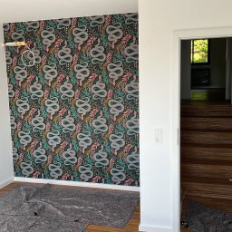 Komfort House Wykończenia Wnętrz Emil Dudziński - Pokój po dekoracji: ściana z wzorzystą tapetą w motywy roślinne i węże, widoczne gniazdka elektryczne, podłoga zabezpieczona folią malarską, otwarte białe drzwi.