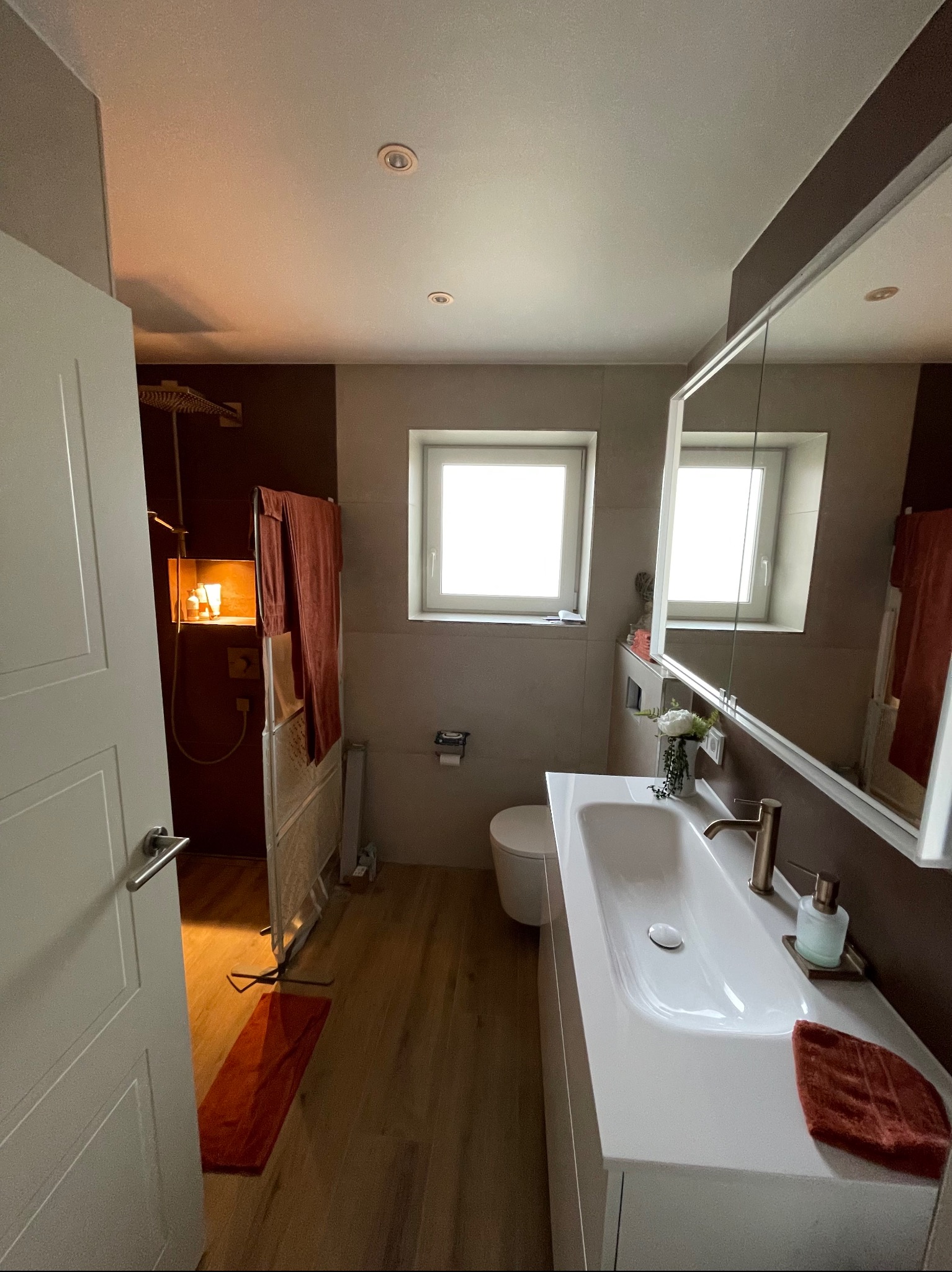 Nowoczesna łazienka z drewnianą podłogą i kabiną prysznicową typu walk-in. Umywalka wpuszczona w blat, lustro z oświetleniem. Widoczne detale wykończenia.