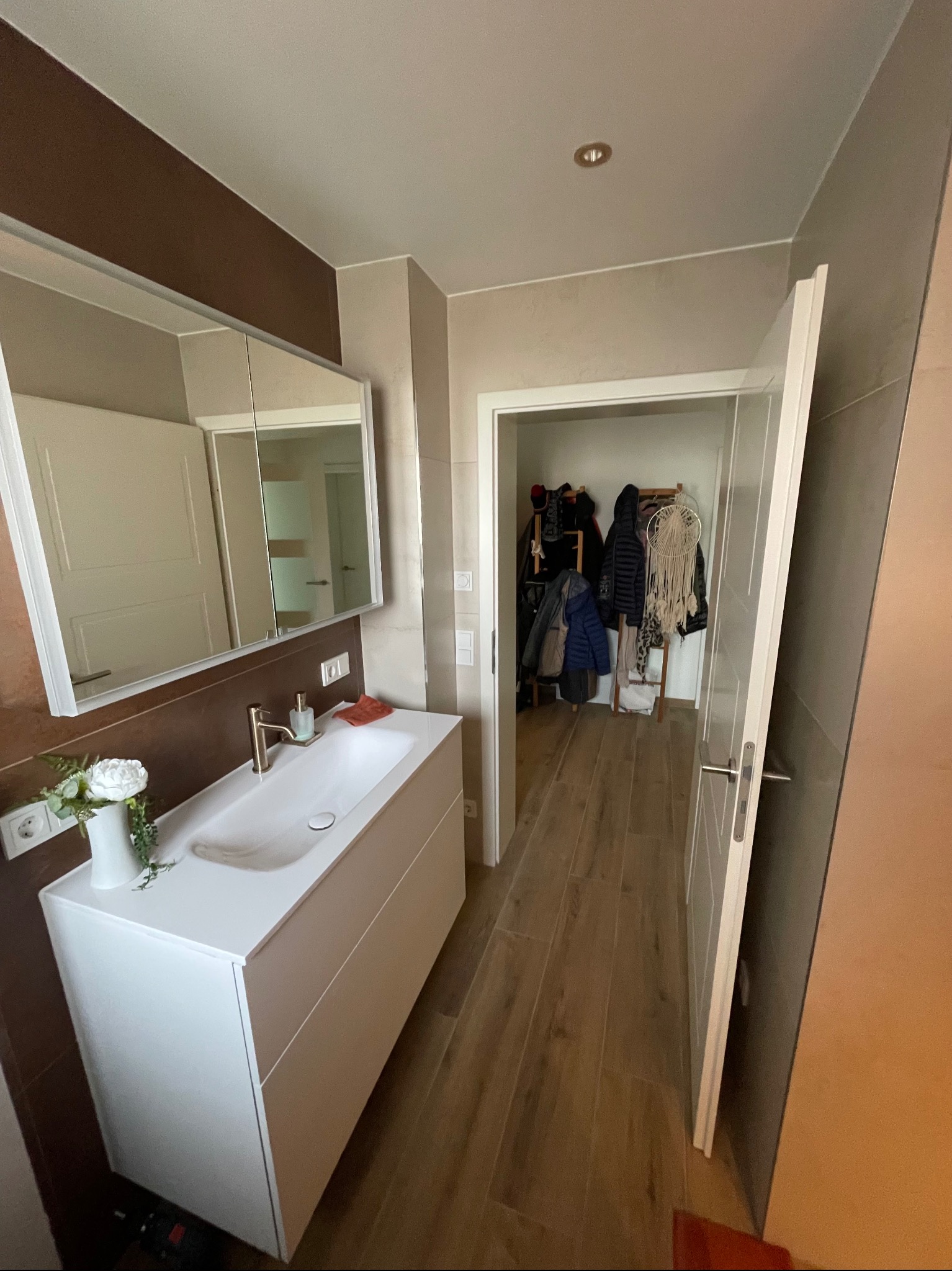 Nowoczesna łazienka po remoncie: biała umywalka z szafką, lustro z brązowym akcentem, drewnopodobna podłoga, otwarte drzwi do garderoby. Minimalistyczny design.