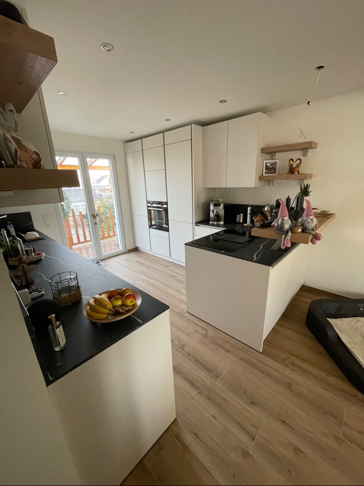 Nowoczesna, minimalistyczna kuchnia z białymi szafkami, czarnym blatem i drewnianą podłogą. Na wyspie kuchennej dekoracje świąteczne. Widok na taras z drewnianym ogrodzeniem.