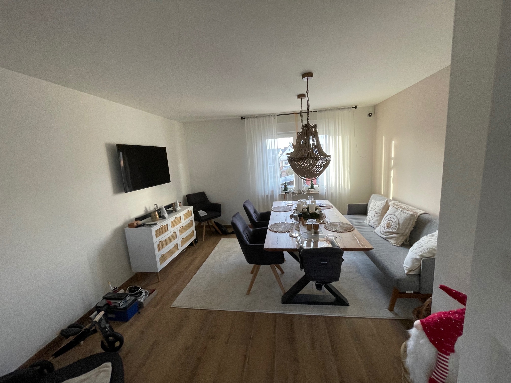 Jasne wnętrze jadalni z drewnianym stołem, krzesłami, sofą i komodą. Na podłodze panele. Centralnie umieszczony ozdobny żyrandol. Minimalistyczny wystrój.