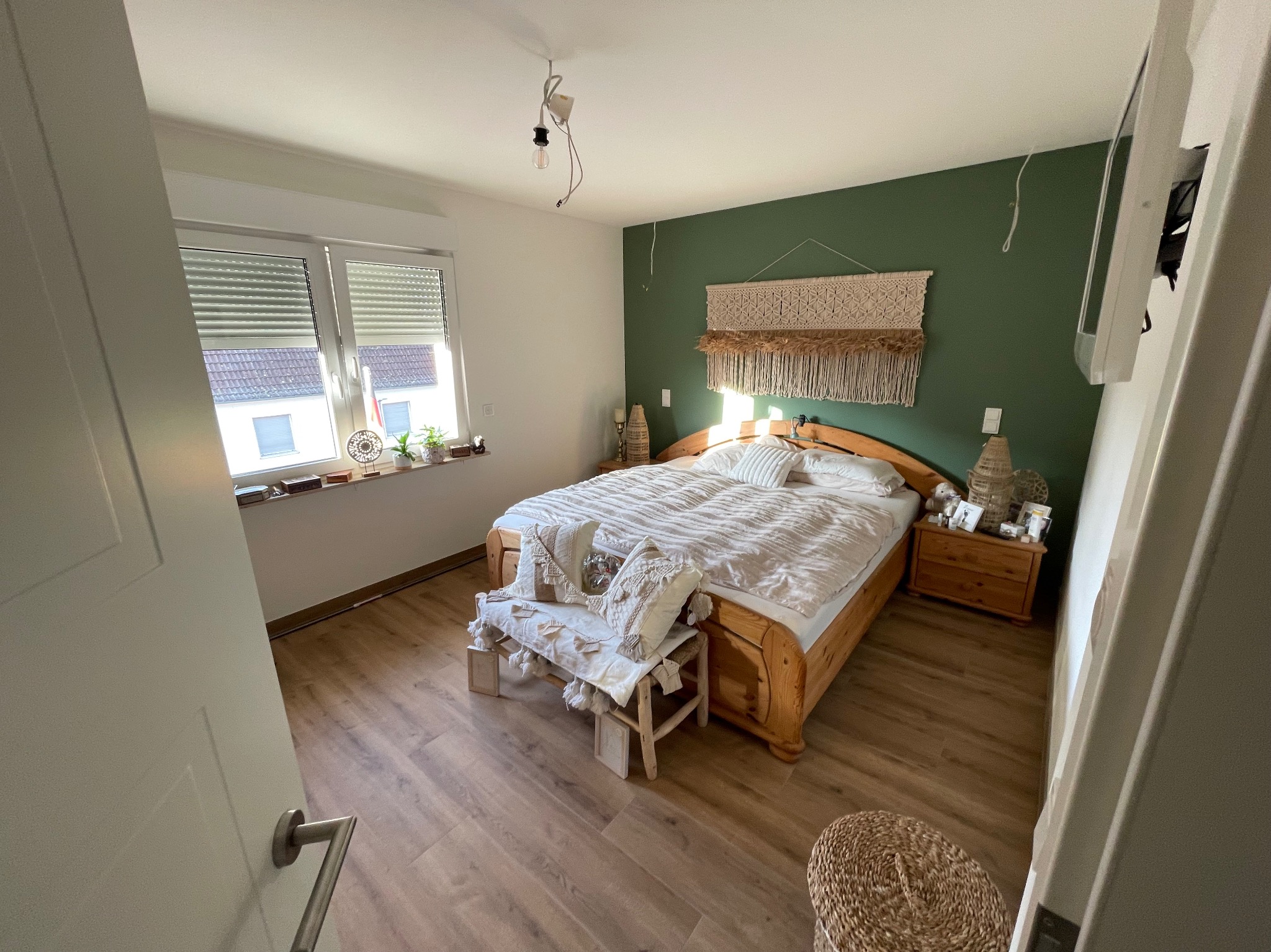 Jasna sypialnia z drewnianym łóżkiem, zieloną ścianą i dekoracyjnym makramem. Widoczna podłoga z paneli i częściowo otwarte drzwi.