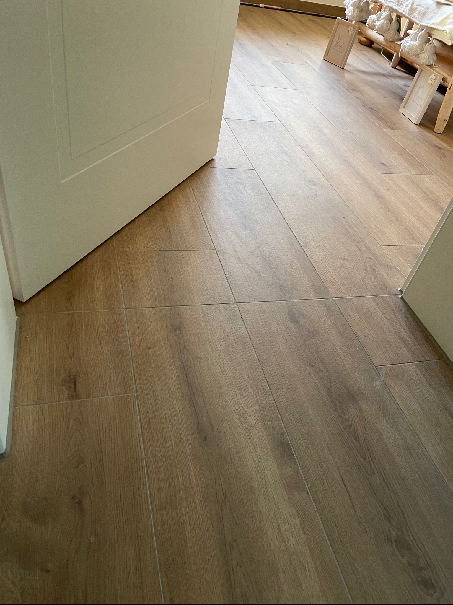 Jasne panele podłogowe ułożone w regularny wzór, widoczne spod uchylonych, białych drzwi. W tle dekoracje i drewniane elementy wystroju.