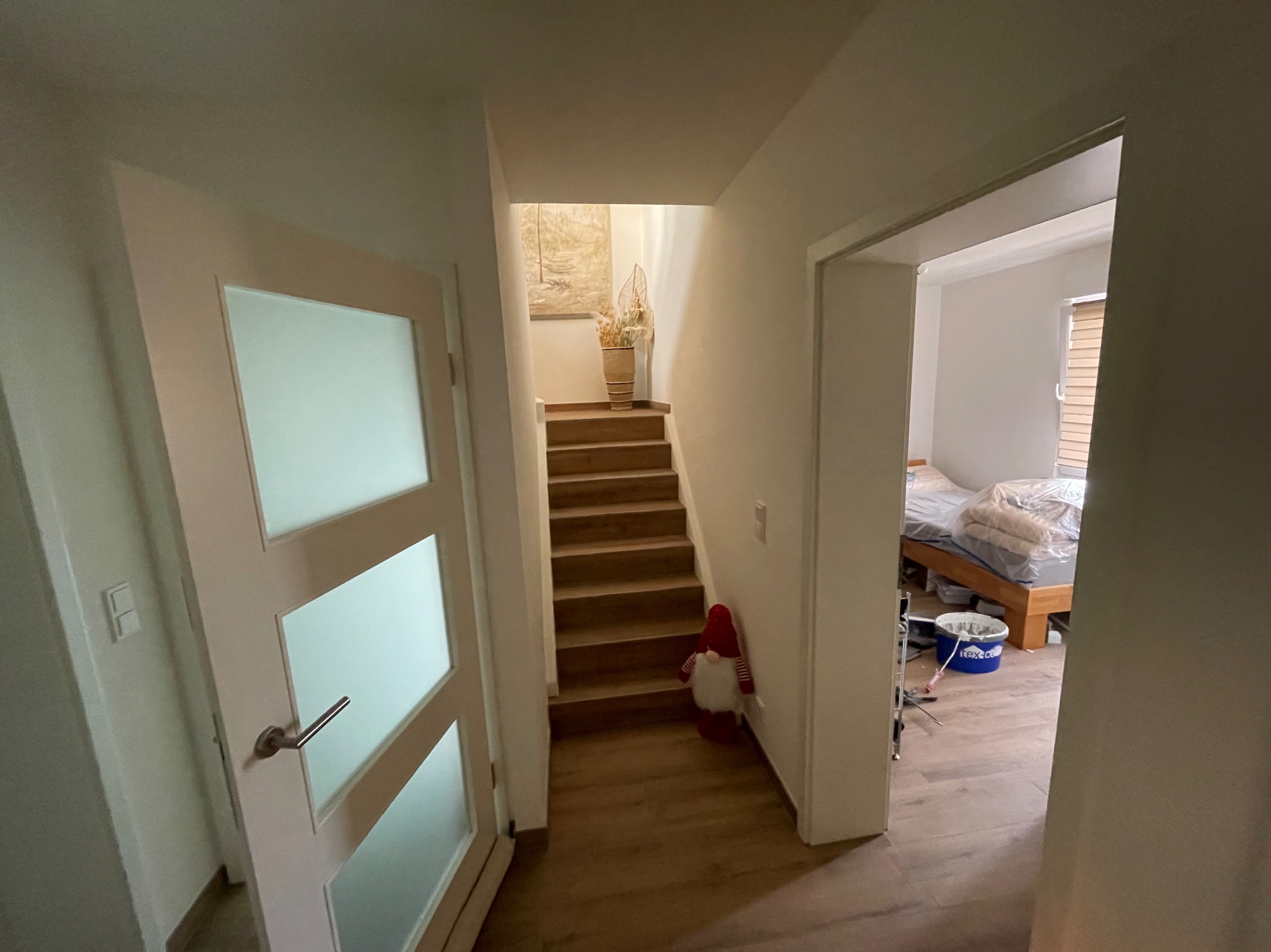 Wnętrze domu po remoncie: widok na schody z dekoracją, drzwi z mlecznym szkłem i fragment sypialni z łóżkiem i wiadrem z farbą. Jasne ściany, drewniane schody i podłoga.