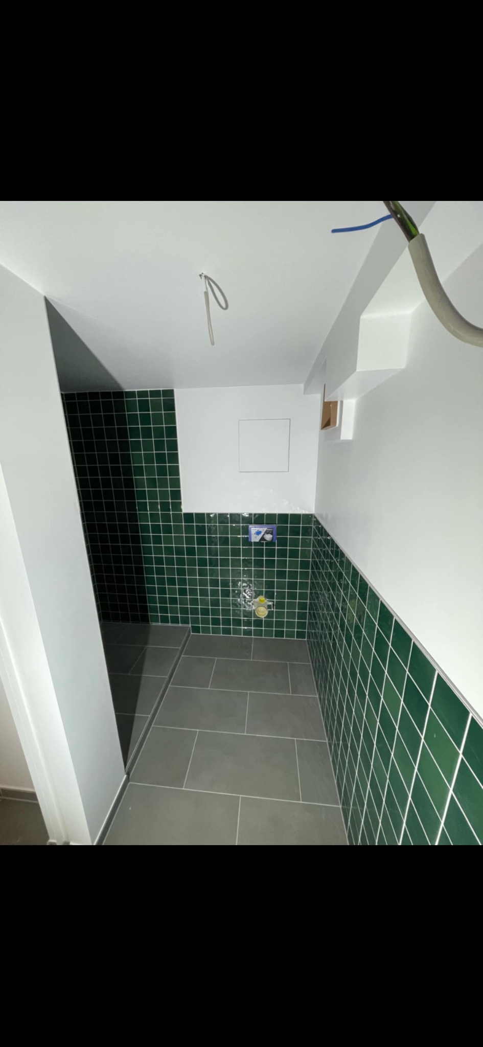 Wykończona łazienka z ciemnozielonymi płytkami ściennymi i szarą podłogą. Widoczne przyłącza wodne, białe ściany i sufit. Minimalistyczny design.