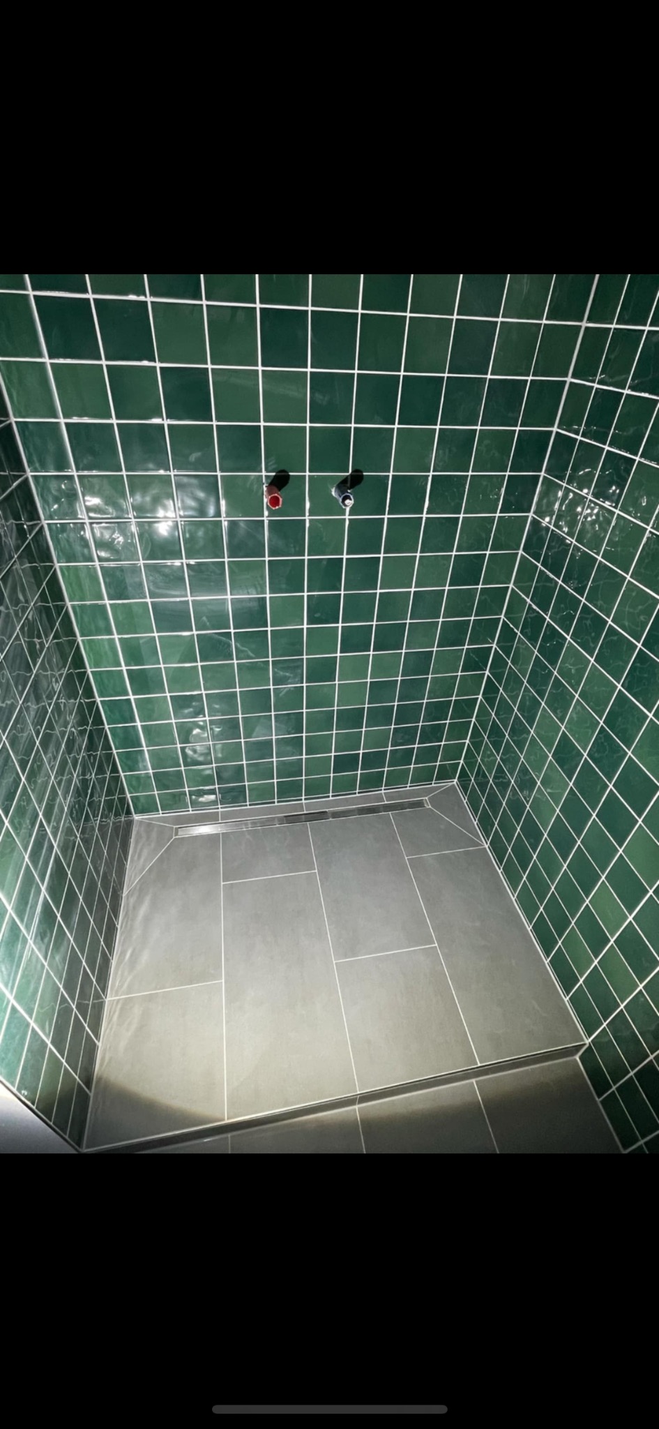 Kabina prysznicowa wyłożona zielonymi, błyszczącymi płytkami z białą fugą i szarą podłogą. Widoczne wystające rury z instalacji wodnej.