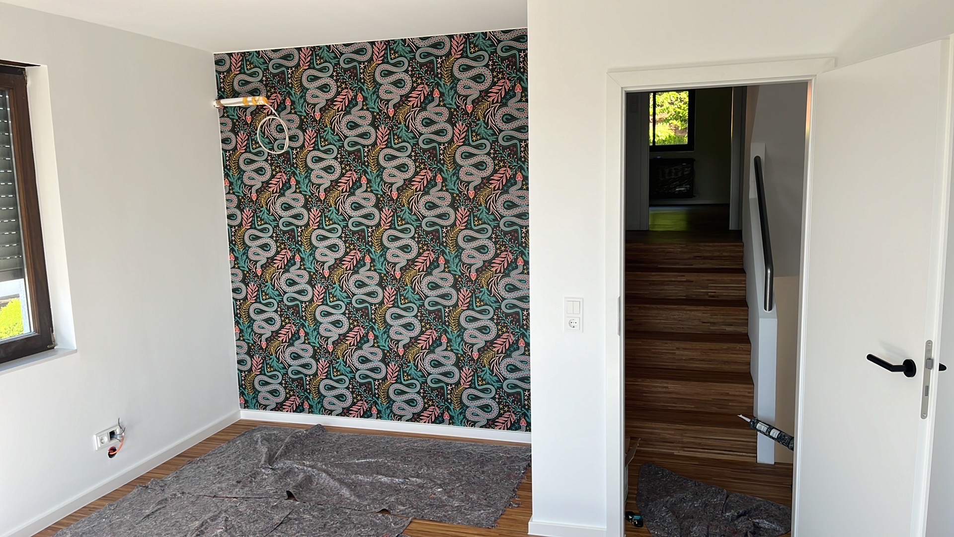 Pokój po dekoracji: ściana z wzorzystą tapetą w motywy roślinne i węże, widoczne gniazdka elektryczne, podłoga zabezpieczona folią malarską, otwarte białe drzwi.
