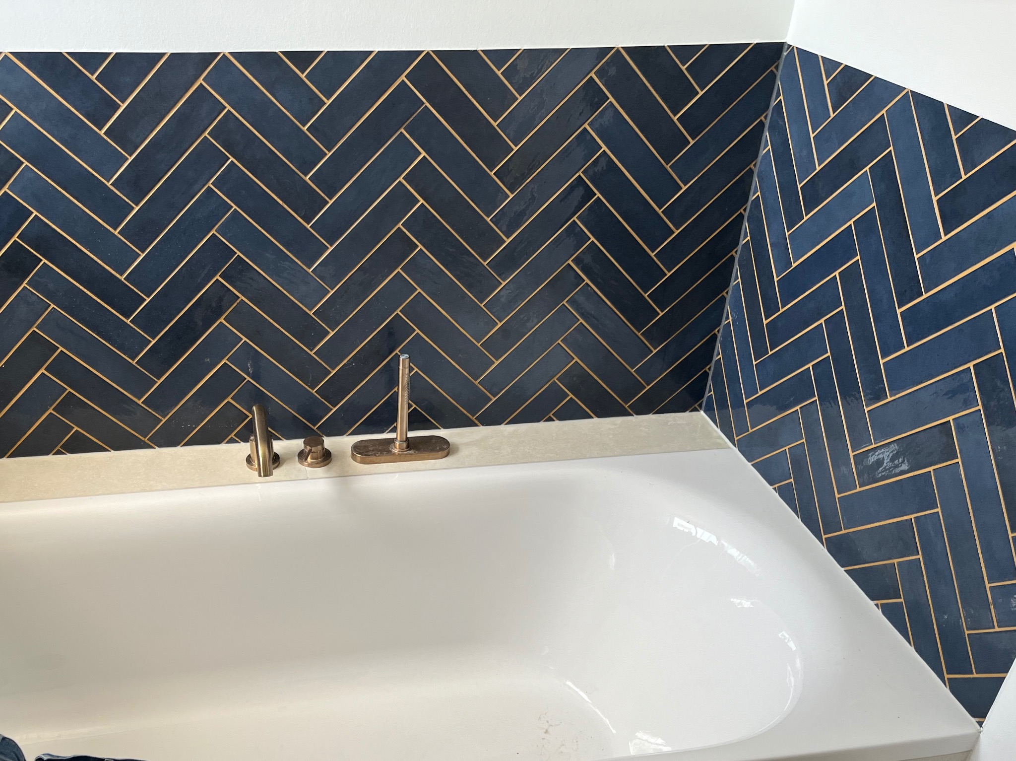 Biała wanna z baterią retro na tle ściany wyłożonej granatowymi płytkami w jodełkę z jasną fugą. Detal wykończenia łazienki.