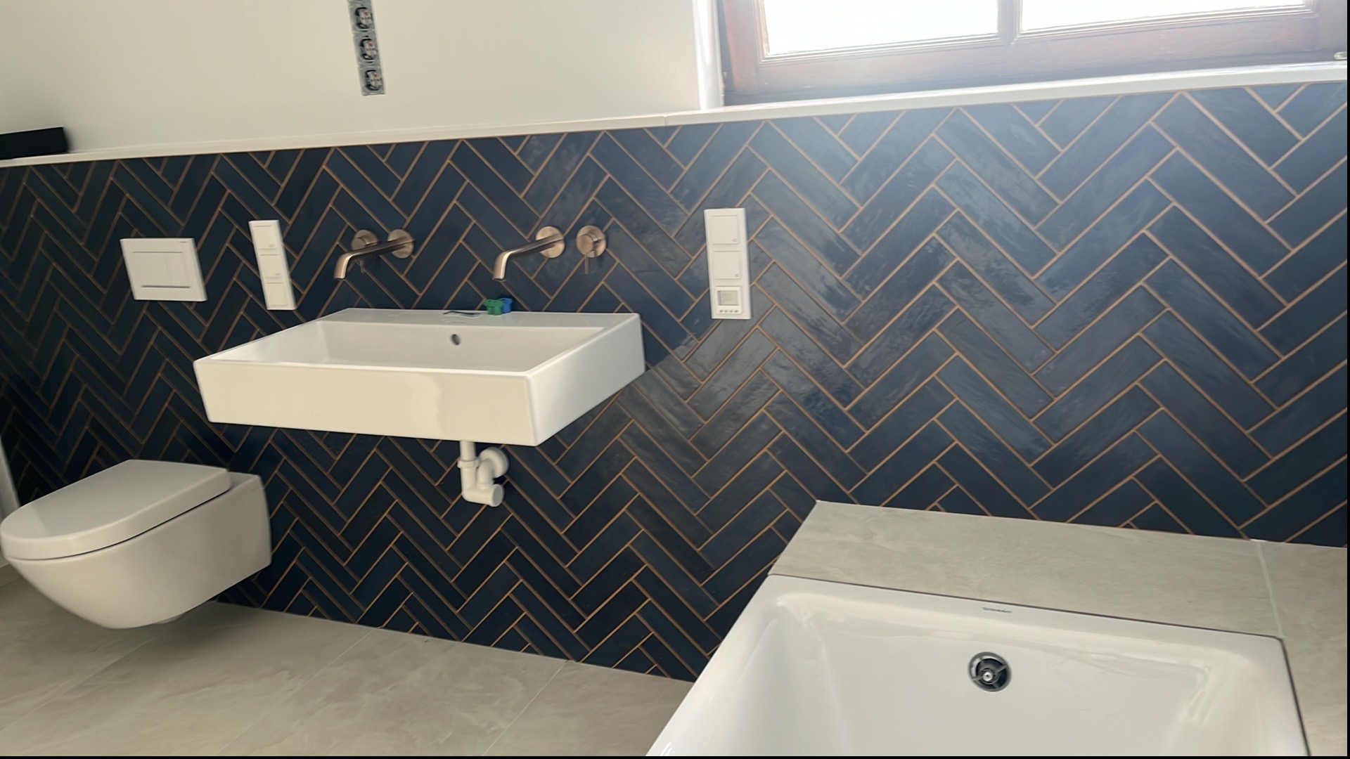 Nowoczesna łazienka z ciemnymi płytkami ułożonymi w jodełkę, białą umywalką, toaletą i wanną. Metalowe baterie w kolorze miedzi. Minimalistyczny design.