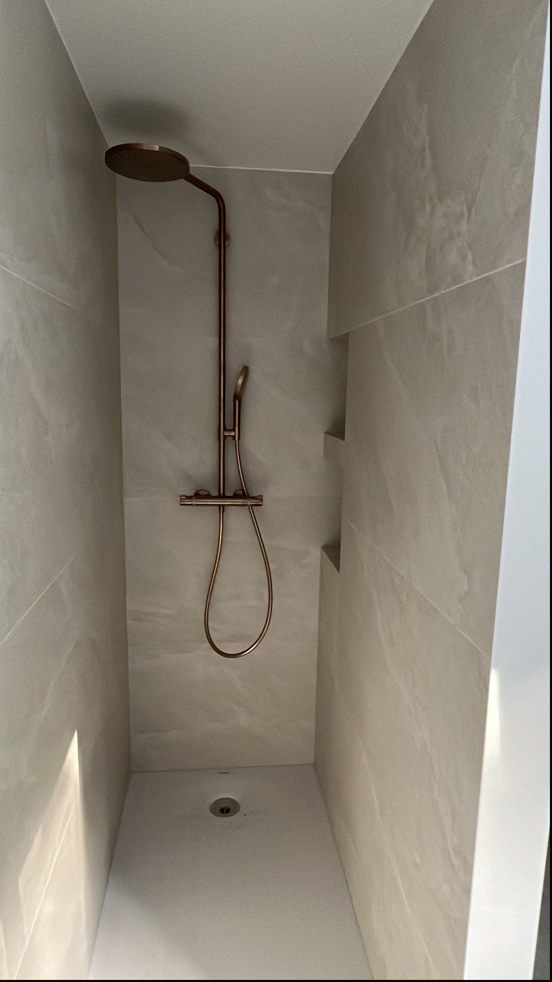 Nowoczesny prysznic walk-in z miedzianą armaturą i półkami w ścianie. Minimalistyczny design, jasne płytki imitujące kamień, odpływ liniowy.