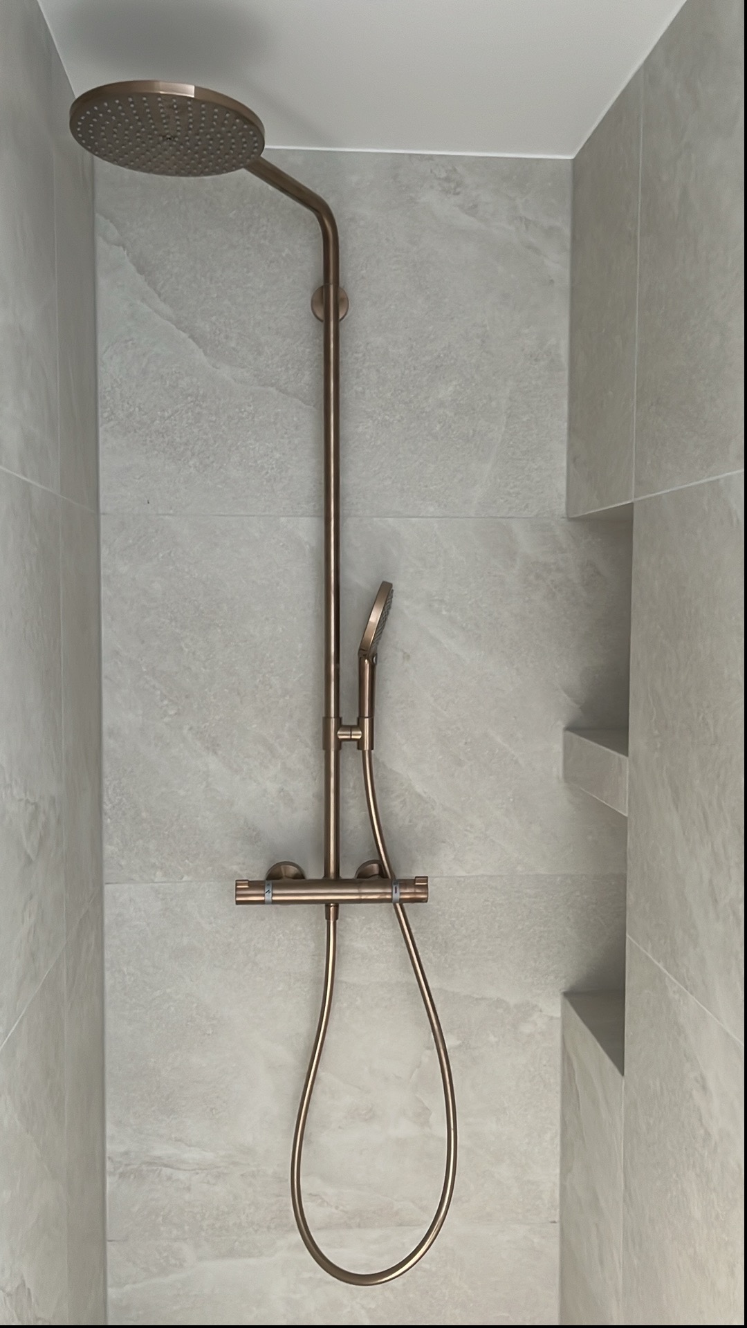 Nowoczesny, miedziany zestaw prysznicowy z deszczownicą i baterią termostatyczną na tle jasnoszarych, marmurkowych płytek w wykończonej łazience.