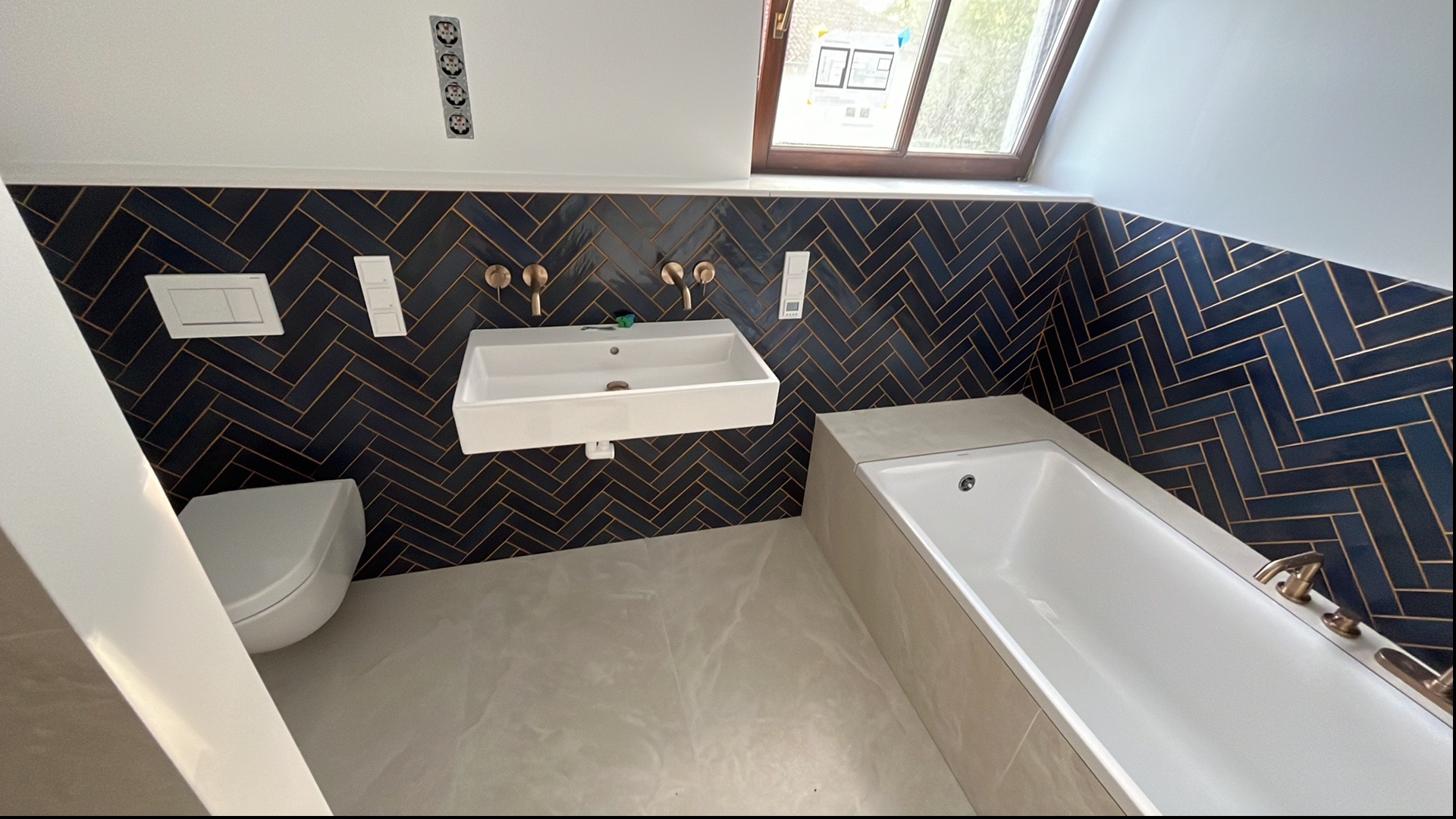 Wykończona łazienka w odcieniach granatu i złota z armaturą w kolorze mosiądzu. Umywalka, toaleta i wanna tworzą spójną całość. Widoczne okno dachowe.
