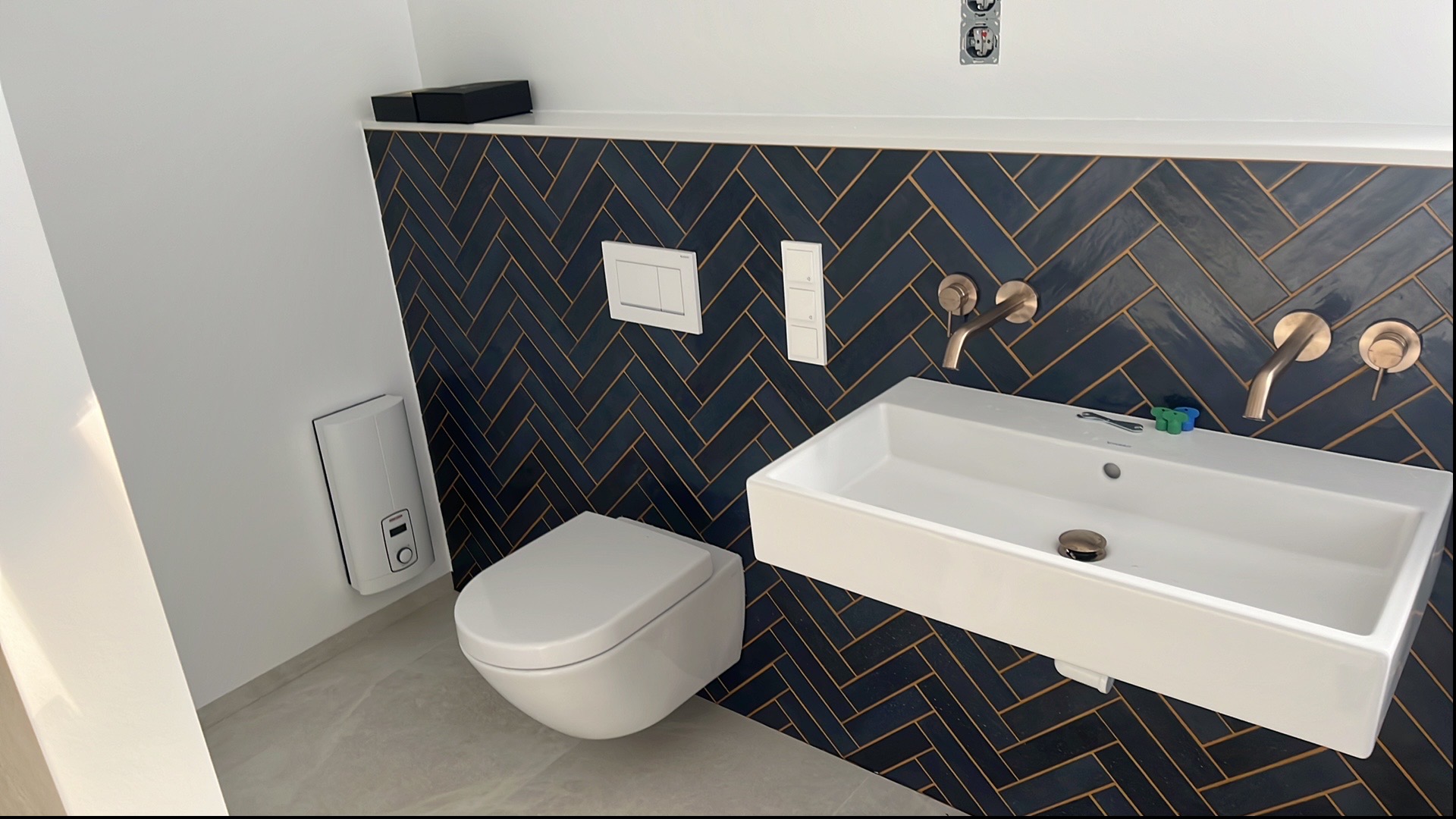 Nowoczesna łazienka po remoncie: ciemne płytki ułożone w jodełkę, biała umywalka i toaleta podwieszana. Widoczny bojler elektryczny i złote baterie.