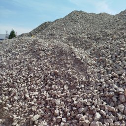 ARTUR JANCZAR AMJ-TRANS - Góra gruzu pod błękitnym niebem. Widoczne różnej wielkości kamienie i fragmenty betonu. W tle zieleń drzew i budynki. Materiał budowlany składowany na placu.