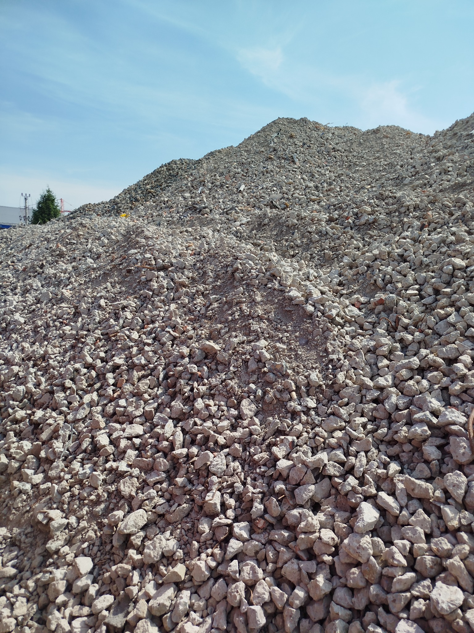Góra gruzu pod błękitnym niebem. Widoczne różnej wielkości kamienie i fragmenty betonu. W tle zieleń drzew i budynki. Materiał budowlany składowany na placu.