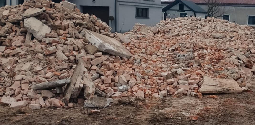 Stos gruzu ceglanego i betonowego na ziemi, z budynkami w tle. Widoczne fragmenty cegieł i betonu w różnych rozmiarach. Ślady opon na ziemi.
