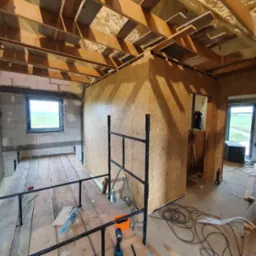 Wnętrze poddasza w trakcie wykończenia: drewniana konstrukcja dachu, ściany z płyt OSB, widoczne ocieplenie pianką, okno z widokiem na zieleń, narzędzia budowlane.