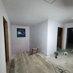 Białe, świeżo pomalowane ściany w pokoju z oknem i otworami drzwiowymi bez ościeżnic. Na podłodze narzędzia malarskie i drabina. Widoczne oświetlenie budowlane.