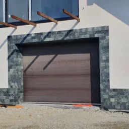 Elewacja domu z garażem, wykończona dekoracyjną okładziną z kamienia naturalnego. Kremowy tynk kontrastuje z grafitową stolarką okienną i bramą garażową.