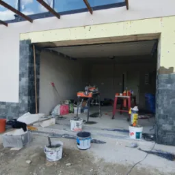 Wnętrze garażu w trakcie wykańczania: widoczne narzędzia, materiały budowlane i fragment elewacji z kamienia dekoracyjnego. Otwór drzwiowy przygotowany do montażu.