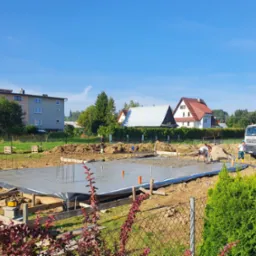 Wylewanie fundamentów pod budowę domu jednorodzinnego, widoczny beton, szalunki i ekipa budowlana przy pracy. W tle domy i błękitne niebo.