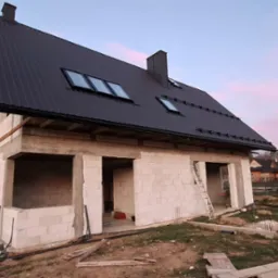 Surowy dom jednorodzinny z czarnym dachem z blachy trapezowej w trakcie budowy, widok z zewnątrz na tle innych dom&oacute;w i drzew.