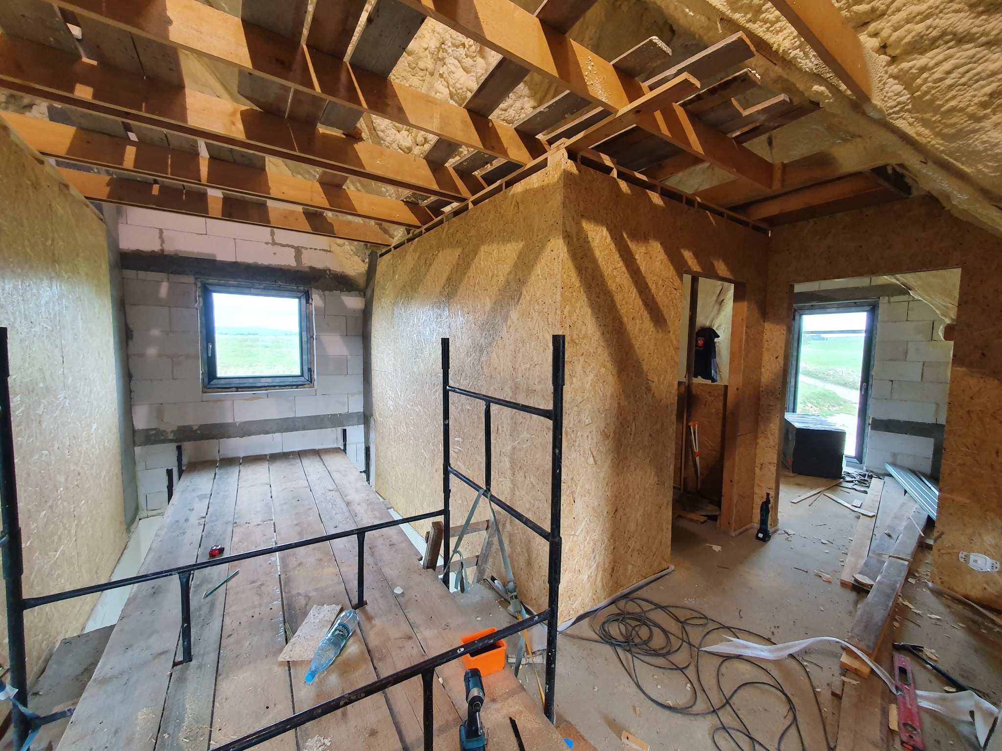 Wnętrze poddasza w trakcie wykończenia: drewniana konstrukcja dachu, ściany z płyt OSB, widoczne ocieplenie pianką, okno z widokiem na zieleń, narzędzia budowlane.