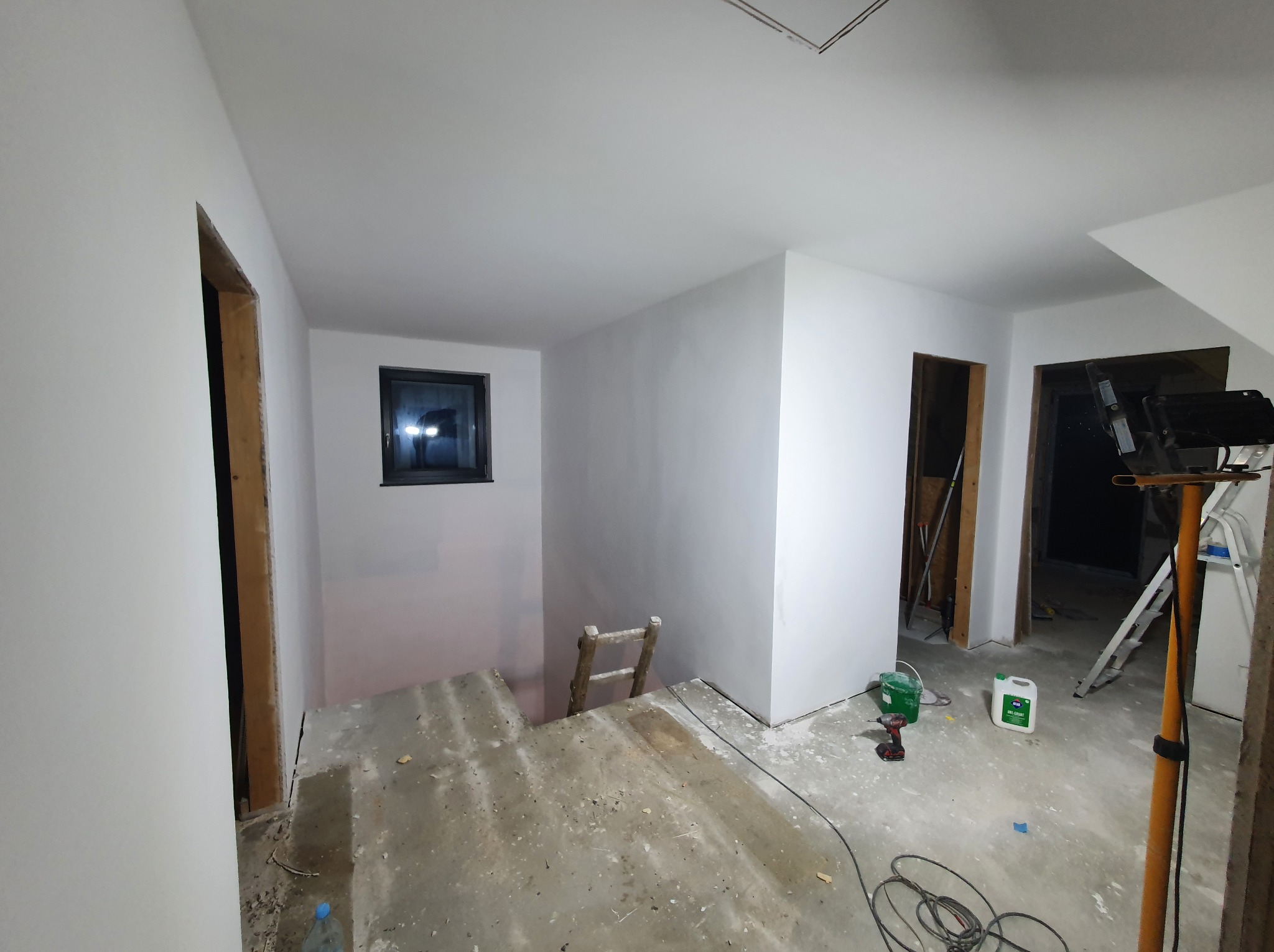 Białe, świeżo pomalowane ściany w pokoju z oknem i otworami drzwiowymi bez ościeżnic. Na podłodze narzędzia malarskie i drabina. Widoczne oświetlenie budowlane.