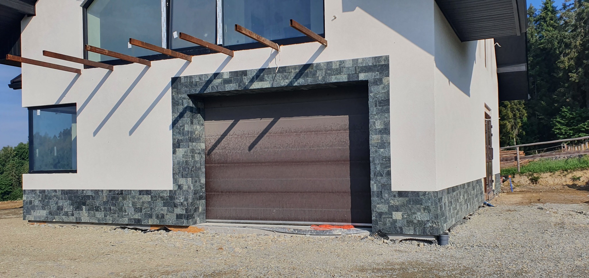 Elewacja domu z garażem, wykończona dekoracyjną okładziną z kamienia naturalnego. Kremowy tynk kontrastuje z grafitową stolarką okienną i bramą garażową.