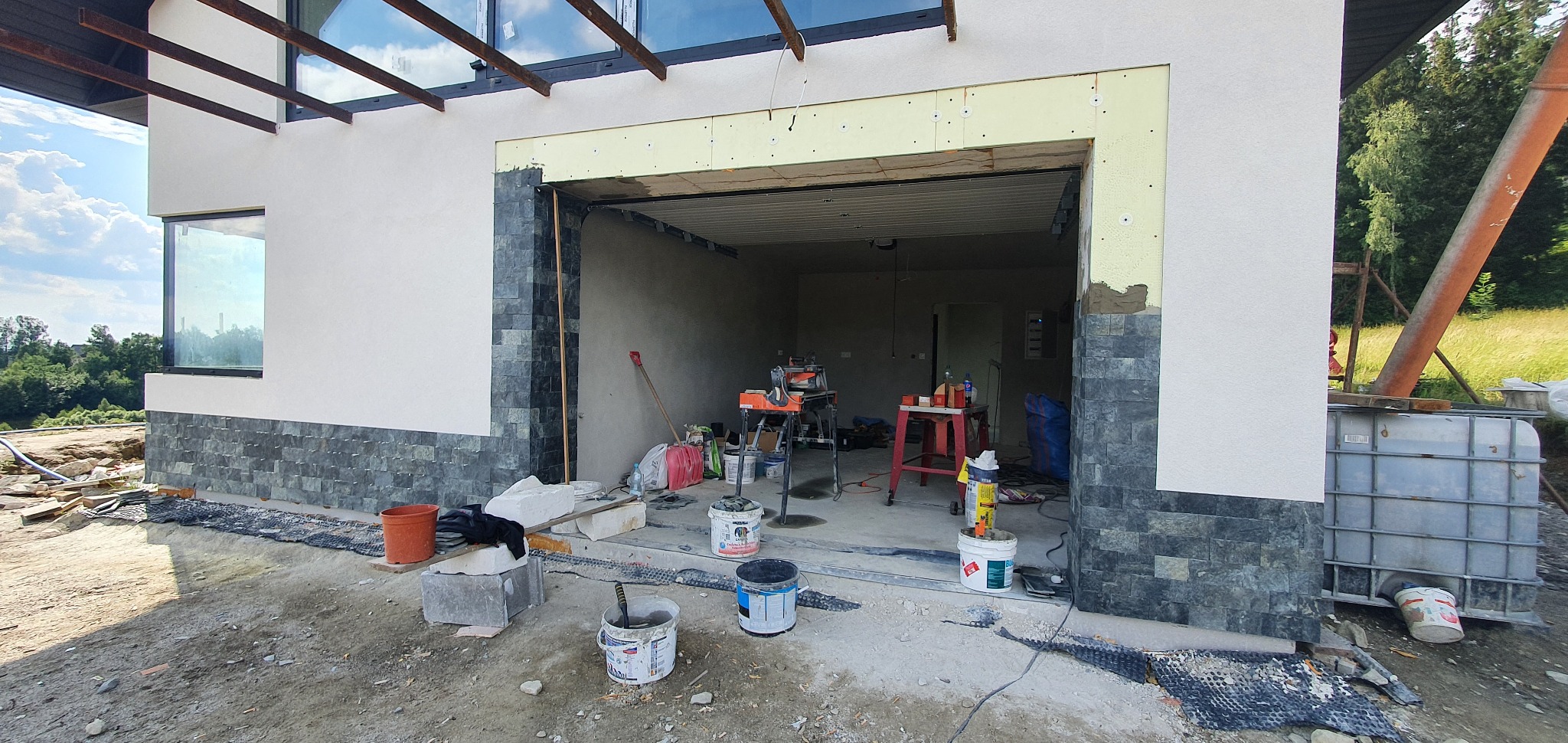 Wnętrze garażu w trakcie wykańczania: widoczne narzędzia, materiały budowlane i fragment elewacji z kamienia dekoracyjnego. Otwór drzwiowy przygotowany do montażu.