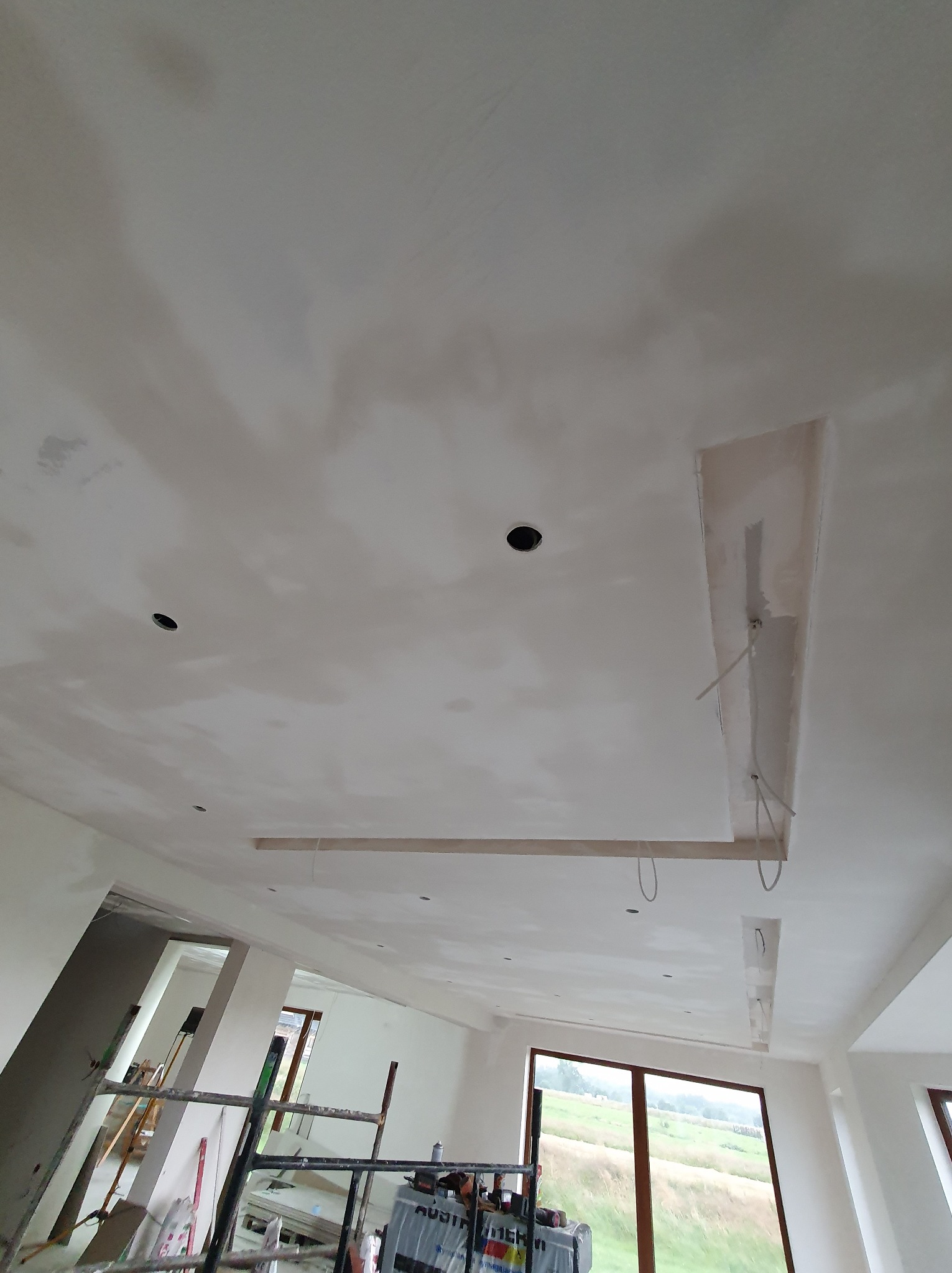 Sufit po szpachlowaniu z widocznymi otworami na oświetlenie i elementami dekoracyjnymi. W tle okno z widokiem na zieleń.