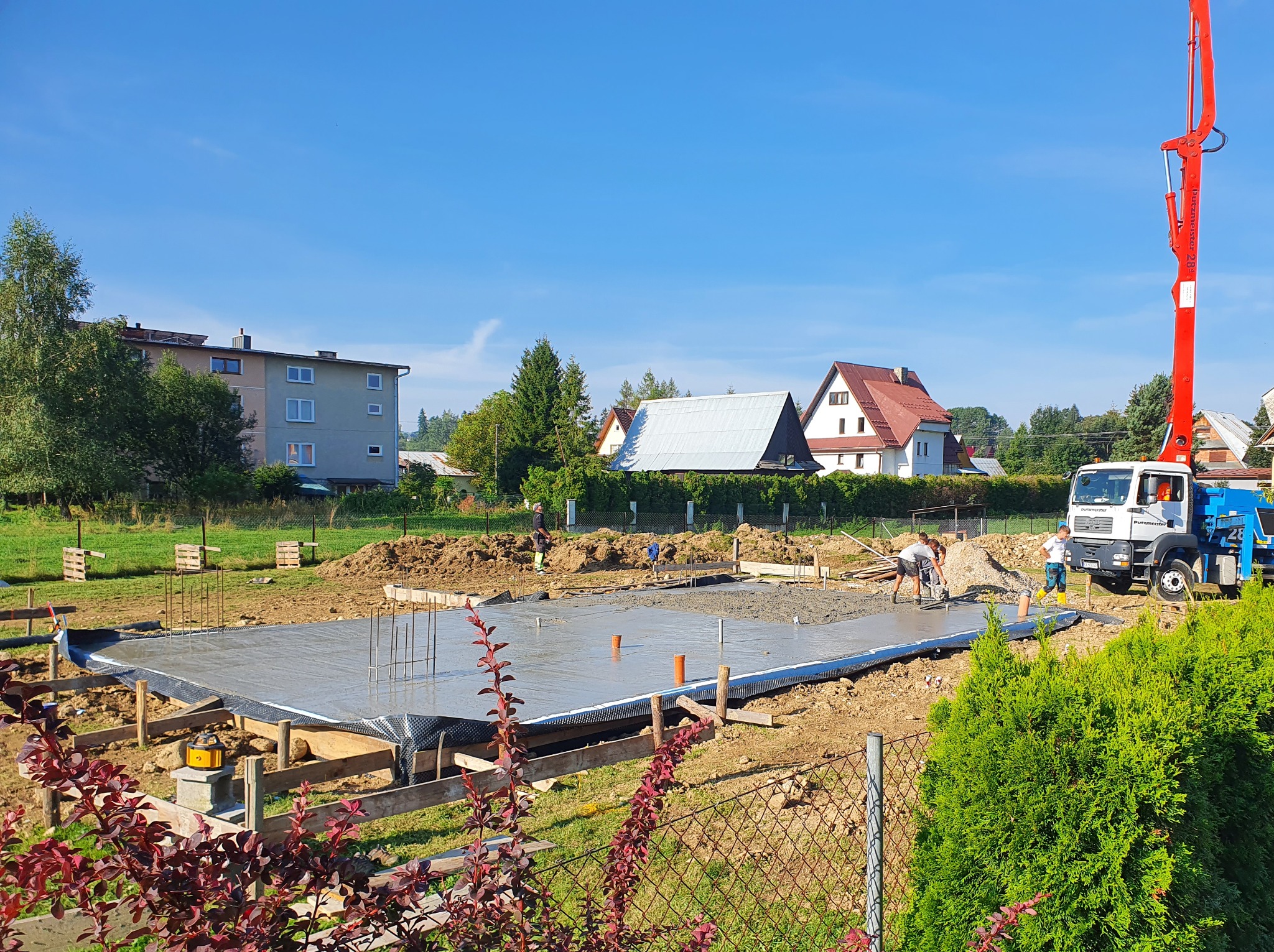 Wylewanie fundamentów pod budowę domu jednorodzinnego, widoczny beton, szalunki i ekipa budowlana przy pracy. W tle domy i błękitne niebo.