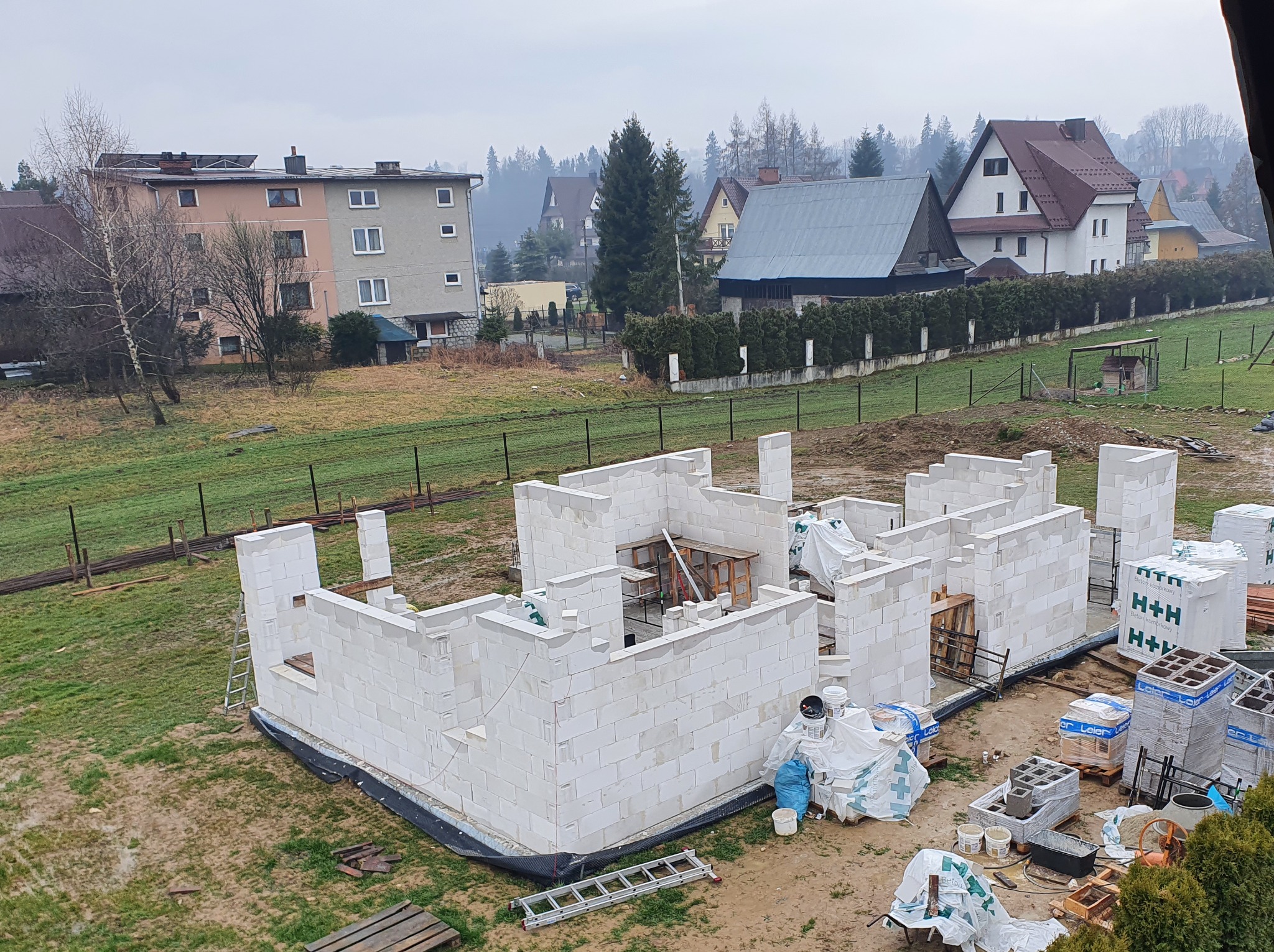 Wznoszenie ścian parteru domu jednorodzinnego z białych bloczków H+H, widok z góry na budowę w otoczeniu zabudowy mieszkalnej i drzew na tle pochmurnego nieba.