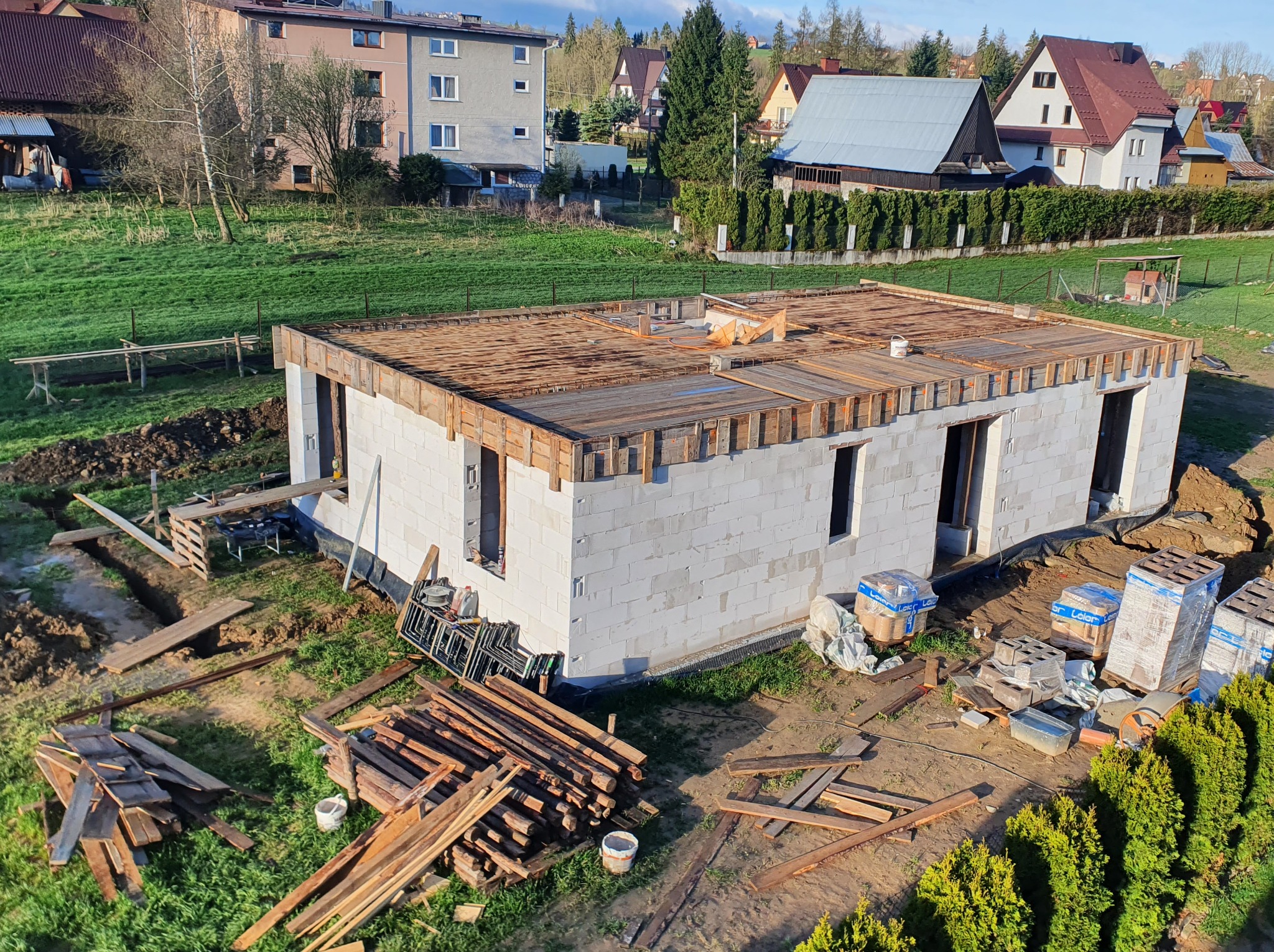Budowa domu jednorodzinnego w stanie surowym otwartym, ściany z białego betonu komórkowego, drewniane szalunki stropu, widok z góry na działkę.