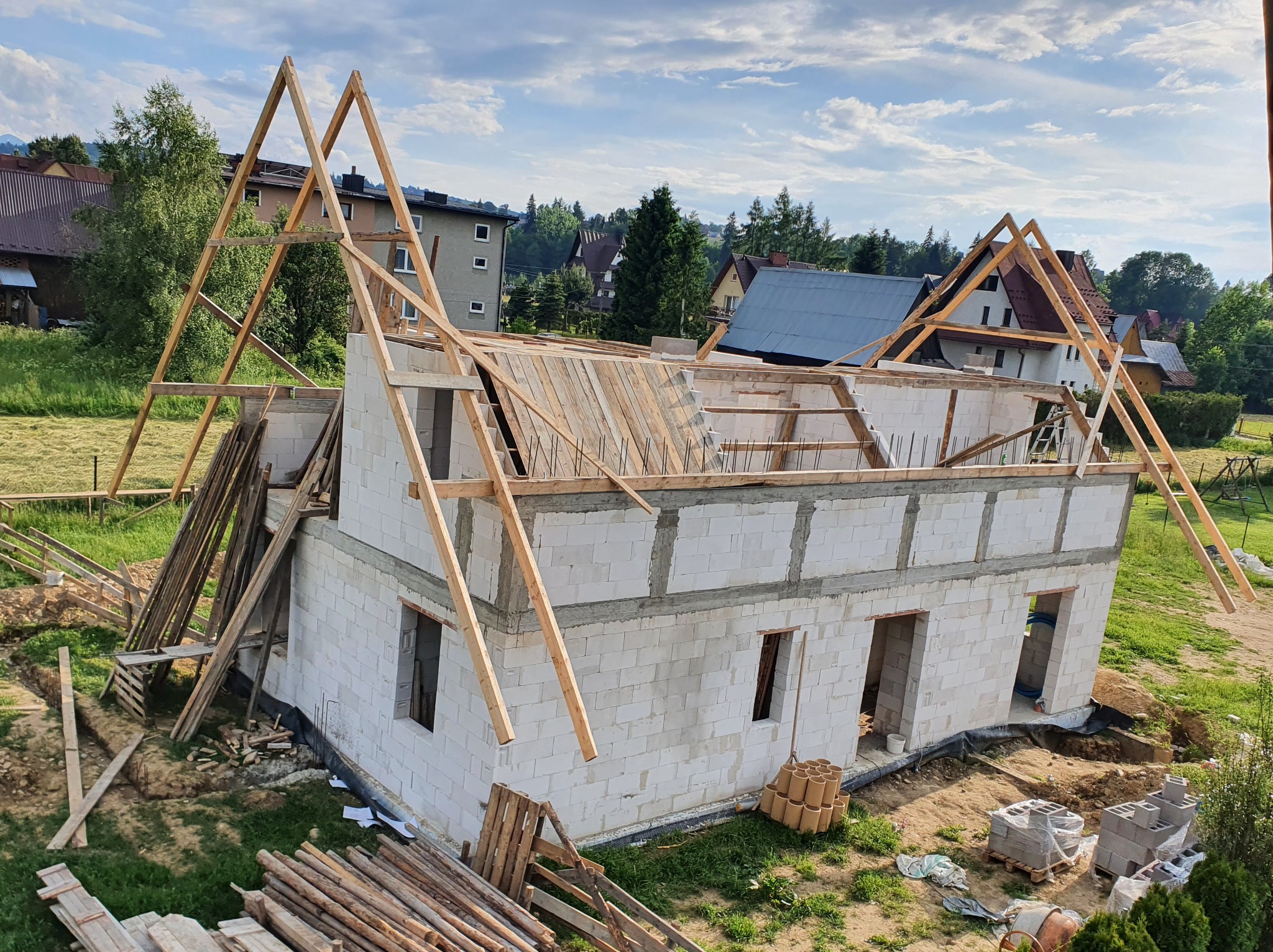 Wznoszenie ścian z białych bloczków i konstrukcji dachu z drewna na budowie domu jednorodzinnego w otoczeniu zieleni i zabudowy mieszkalnej.