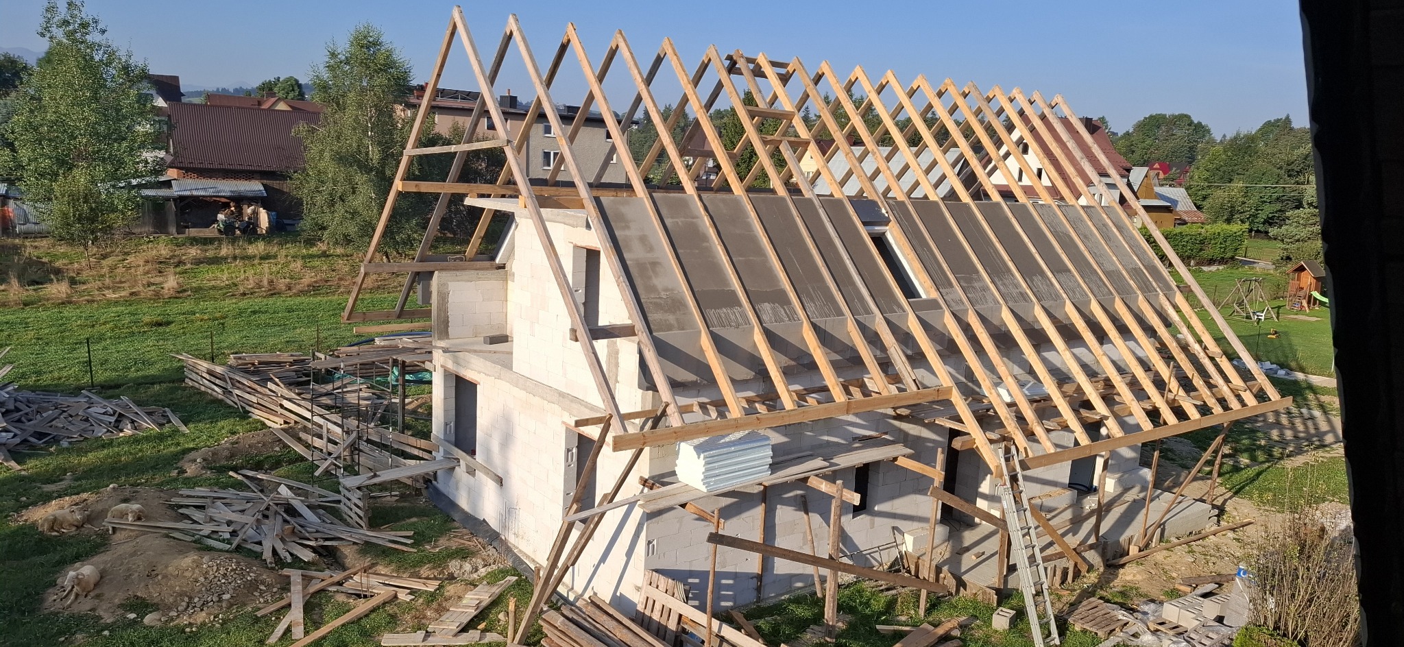 Budowa domu jednorodzinnego: wznoszenie drewnianej konstrukcji dachu na murowanych ścianach z betonu komórkowego. Widoczne elementy izolacji. Krajobraz wiejski w tle.