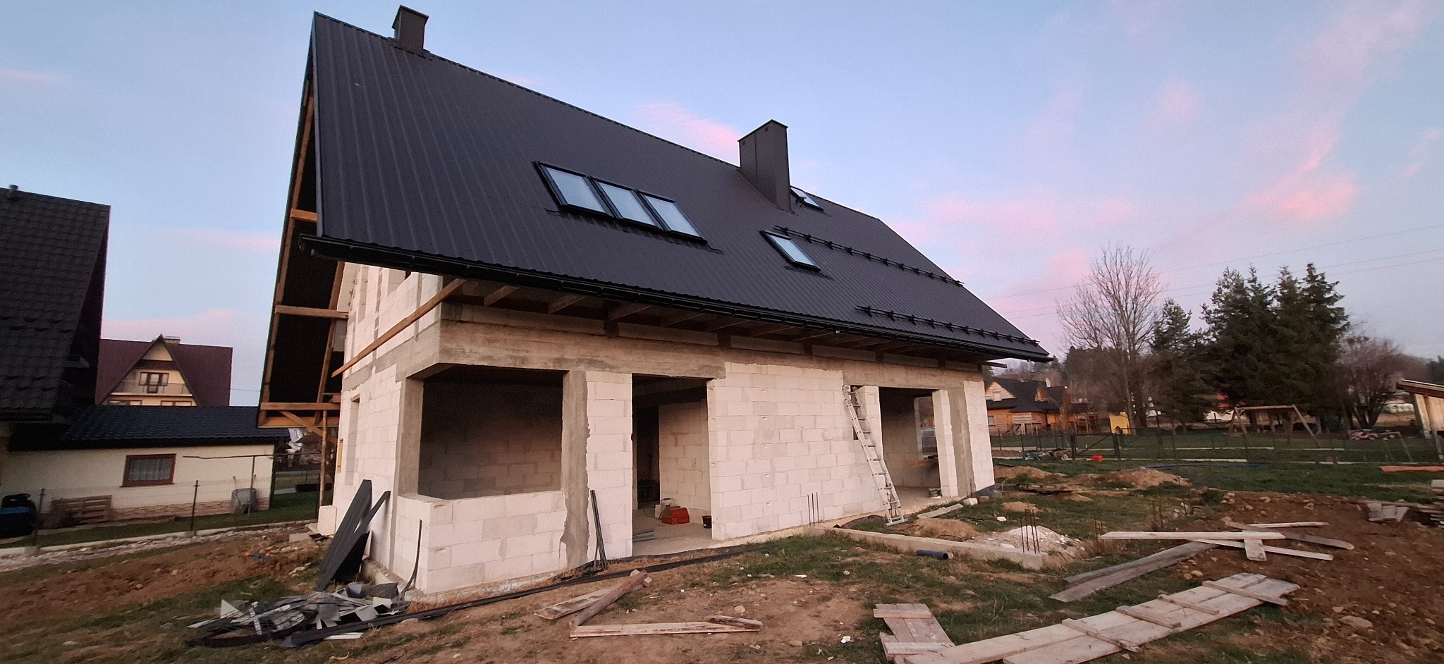 Surowy dom jednorodzinny z czarnym dachem z blachy trapezowej w trakcie budowy, widok z zewnątrz na tle innych domów i drzew.