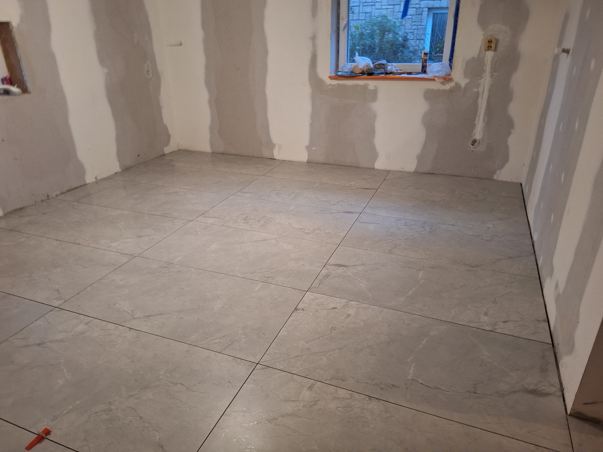 Ułożona szara podłoga z płytek w pomieszczeniu w trakcie remontu, widoczne ślady po pracach na ścianach i okno z widokiem na zewnątrz.