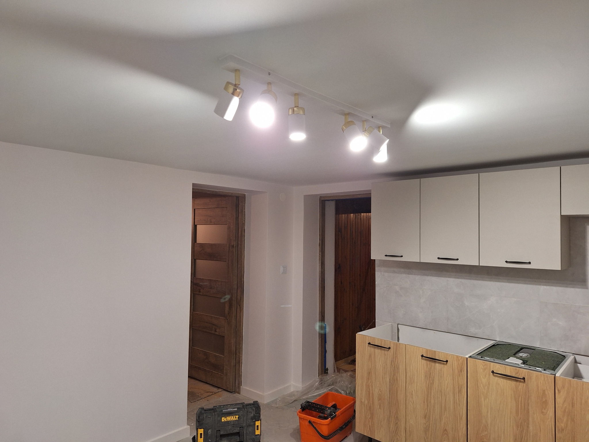 Wnętrze w trakcie prac wykończeniowych: widoczna ściana po przygotowaniu do malowania, zamontowane oświetlenie punktowe, zabudowa kuchenna bez blatu, narzędzia malarskie w tle.